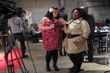 Two women being filmed, one speaking into a handheld microphone, at a clothing event.