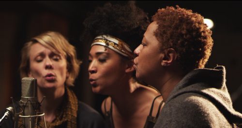 Three backing singers gathered close around a studio microphone, mid-phrase.