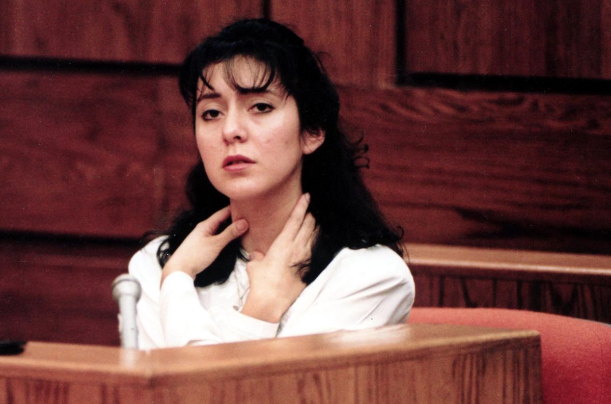A dark-haired woman on a witness stand in a wood-panelled courtroom, one hand at her throat, speaking into a microphone.