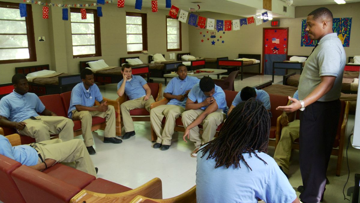 Young men in matching blue shirts sit in a circle in a facility common room hung with bunting, while an adult stands addressing them.