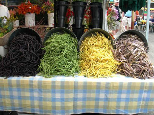 A farmers' market table under a blue and yellow checked cloth, with wide bowls heaped with green, yellow and dark beans, and cut flowers behind.