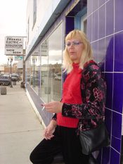 A woman in a red top and patterned jacket standing on a pavement beside a purple tiled shopfront.