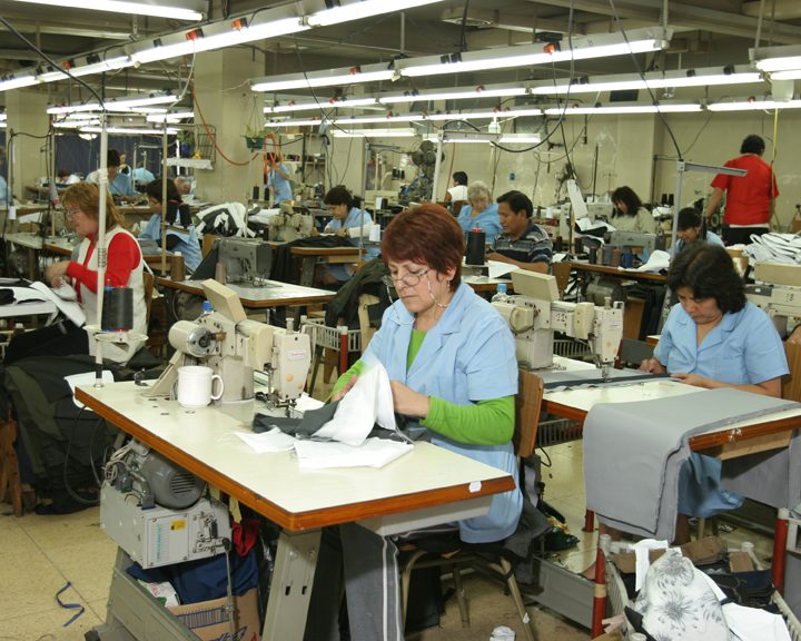 A garment factory floor under fluorescent lights, with rows of sewing machines and workers, mostly women, at their stations.