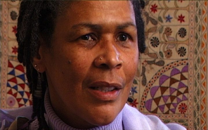 Close-up of a woman with locs speaking, a patterned quilt hanging behind her.