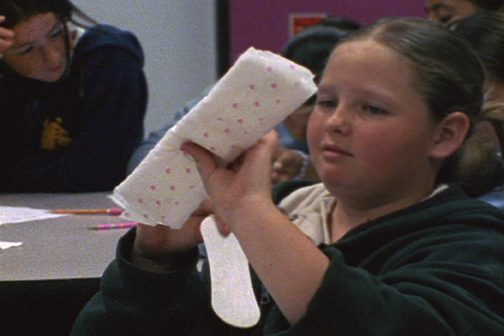 A girl in a classroom holding up a sanitary pad and its wrapper, other students seated behind her.