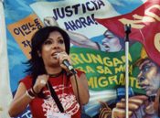 A woman speaking into a microphone in front of a painted banner reading Justicia Ahora.