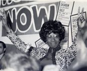 Black-and-white photograph of a woman with an afro speaking amid a crowd, campaign placards raised behind her, one of them reading NOW.